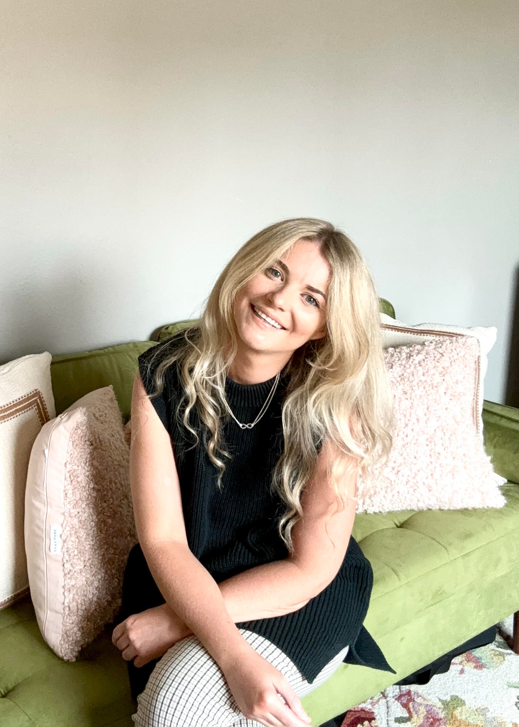 Hannah Spivak sitting on a green velvet sofa with blush and cream cushions, smiling toward the camera, wearing a black sleeveless knit top and a silver necklace with an infinity pendant.