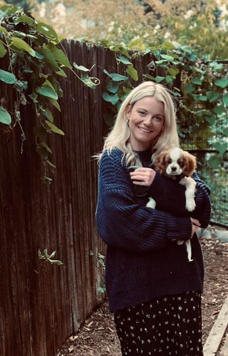 Picture of Hannah, a white woman with blonde hair, wearing a blue sweater, holding her dog, standing outside in front of a wood fence.
