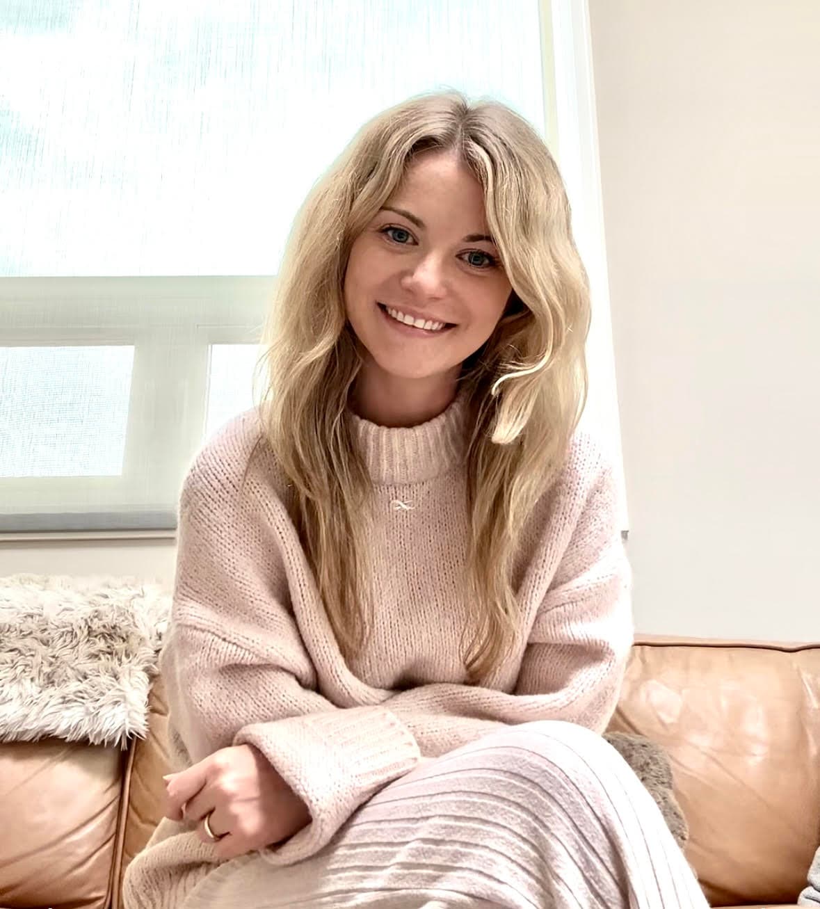 Hannah Spivak, a white woman with long blonde hair wearing a soft pink sweater, sitting on a couch by a bright window and smiling.